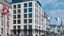 Modern white building with glass windows, multiple floors, and a blue sign reading 'Z ZURICH', located in a busy city street with people walking and streetlights.