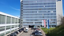 multistory building, numerous glass windows, open parking lot, Swiss flags, white and blue exterior