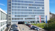 A large multi-story office building with a parking lot in the foreground. The building has a modern, rectangular design with many windows. The parking lot appears to have several cars parked in it.