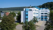 modern multi-story building, white exterior, blue accents, parking lot, scenic mountain view, several windows, entrance with steps