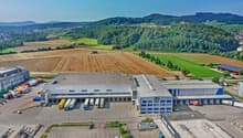 Large industrial building with multiple loading docks, surrounded by parking lots, trucks, and a vast green landscape.