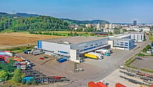 Large industrial building with multiple trucks parked in front, surrounded by a parking lot and a field.