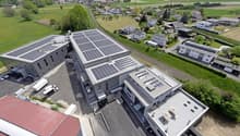 Large building with solar panels on the roof, multiple parking spaces, surrounded by houses and greenery