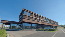 modern office building, glass facade, spacious parking area, brown facade, cars parked in front, trees and bushes, sign with 'Reisezentrum'