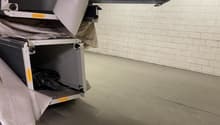 The image shows a storage room or utility area with several large storage containers or bins stacked on shelves. The room has a concrete floor and exposed ceiling beams, indicating an industrial or utilitarian space.