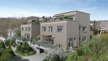 3-story apartment building with gray exterior, large windows, balconies, and a landscaped garden area with trees and shrubs in the foreground