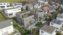 Multi-story apartment building with balconies, raised ground floor, and parking spaces. The building has a modern, minimalist design with a mix of white and gray colors. The surrounding area appears to be a residential neighborhood with other houses and b