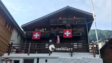 2-story wooden chalet-style building with balconies, Swiss flags displayed, and a sign for the 'Restaurant Obermatt'