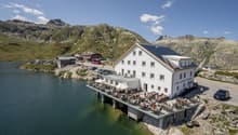 A white hotel building with a terrace overlooking a scenic mountain lake surrounded by rocky peaks. There are people dining and relaxing on the terrace. The hotel appears to be situated on a cliff or elevated platform with a parking area below.
