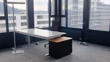 This image shows a modern office space with large windows providing a view of the surrounding cityscape. The room has a minimalist design with a white table, a black office chair, and a gray carpeted floor. The ceiling features a grid-like pattern of pane