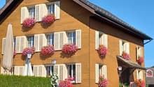 2 story house, brown wooden exterior, white shutters, pink flowers in window boxes, garden with hedges and plants, awning over entrance
