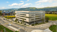 Office building with multiple floors, glass windows, located near a road intersection, surrounded by greenery and mountains in the background.