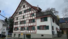 white and brown house, multiple windows, red and green shutters, brick chimney, balcony on top floor