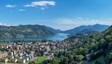 Scenic view of a lakeside town surrounded by mountains and lush greenery. The image shows a picturesque landscape with a large body of water, likely a lake, surrounded by hills and mountains. The town is situated along the shoreline, with colorful buildin