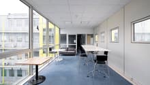 Large open office space with floor-to-ceiling windows, gray carpeted floors, white walls, and modern furniture including a sofa, tables, and chairs. The space appears to be designed for a professional work environment.