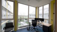 Spacious office with large windows providing a view of the outdoor scenery, including trees and buildings. The office is furnished with a desk, computer, and office chairs. The walls are painted in a light color scheme, and the floor is covered with blue