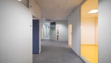 Empty hallway with gray carpeted floor, white walls, ceiling with lights, blue and green doors, and fire extinguisher