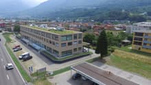 Aerial view of a building with a green roof, surrounded by a parking lot with several cars parked, a road with cars driving, and a mountain in the background.