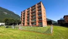 large orange building, many balconies, large red square outlined, tall green trees, grassy ground, mountains in background