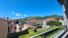 View from a balcony with a railing, overlooking a street with buildings, mountains, a car, and a tree.