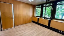 The image shows a spacious office room with wooden paneled walls, a wooden floor, and large windows overlooking a green outdoor area. The room has a clean and modern design, with built-in storage units along the walls.
