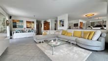 Spacious open-concept living room with large sectional sofa, glass coffee table, and artwork on the walls. The room has a modern and bright design with tile flooring and recessed lighting.
