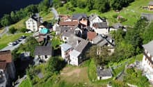 Aerial view of a small village nestled in a hilly, forested area. The village consists of traditional stone and tile-roofed buildings, some raised off the ground, with narrow streets and paths winding through the landscape.