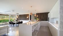 modern interior, white countertops, open-plan, large windows, large dining table, 2 stools, 2 couches, wooden wall, chandeliers