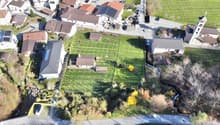 Aerial view of a residential area with several houses, a church, and a small stream. The houses are surrounded by green fields and trees. Some of the houses have solar panels on their roofs.