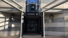 Modern building entrance with glass doors, metal frame, and a sign reading 'LISANO 3'. The entrance is flanked by two lamps.
