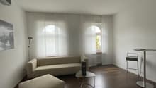 large sofa, foot stool, round table, floor lamp, two large arched windows with curtains, picture frame, hardwood floor, ceiling lamp, white walls
