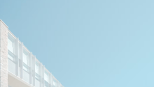 The image shows a modern, minimalist building with a series of large, rectangular windows arranged in a grid-like pattern. The building has a clean, white exterior and is set against a clear blue sky, creating a simple and elegant visual composition.
