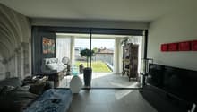 Spacious living room with large sliding glass doors leading to a balcony or terrace, hardwood floors, modern furniture including a sofa and armchair, decorative artwork on the walls, and a shelving unit with various decor items.