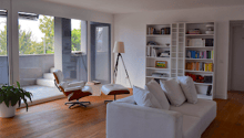 Living room, white walls, hardwood floor, glass door to terrace, white couch, armchair, books on shelf, lamp, plants