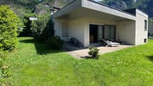 Single-story modern house with a flat roof, surrounded by lush greenery and mountains in the background. The house has large windows and a balcony or terrace area.