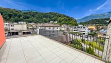 Spacious balcony with tiled flooring, metal railing, overlooking a residential area with buildings, trees, and mountains in the background.