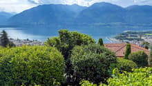 The image shows a scenic view of a mountainous landscape with a large lake in the foreground. The mountains are surrounded by lush, green vegetation, and the lake is dotted with boats and other watercraft. The city can be seen in the distance, with red-ti