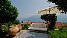 The image shows an outdoor patio area with a comfortable sofa, a large umbrella, and two large terracotta pots. The patio overlooks a scenic landscape with mountains in the background, suggesting a beautiful view. The patio is surrounded by lush greenery,