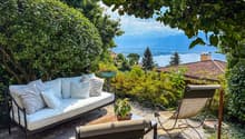 Cozy outdoor patio with a comfortable sofa, chairs, and a scenic view of the lake and mountains in the background, surrounded by lush greenery and trees