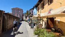 Scenic narrow street with buildings on both sides, multi-story buildings, parked bicycles, a cyclist, awnings, potted plants, tables, chairs, bicycles, mountains in the distance