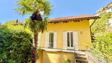 Two-story house, yellow exterior, red tile roof, balcony on the first floor, windows with white shutters, steps leading up to the entrance, front yard with palm tree and greenery.