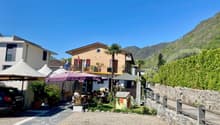 A building with two floors, beige exterior, outdoor dining tables, plants, flags, a small house, mountains, and a clear sky.