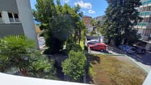 View of a parking area with several cars parked, surrounded by trees and bushes, with a building in the background.