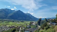 The image shows a scenic mountain landscape with a village nestled in the valley below. The mountains in the background are tall and rugged, with a blue sky dotted with clouds overhead. The village consists of a cluster of buildings with red-tiled roofs,