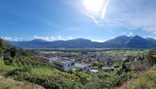 The image shows a panoramic view of a town or city surrounded by mountains. The town has a mix of residential and commercial buildings, with a modern-looking building visible in the foreground. The landscape is lush and green, with trees and vegetation co