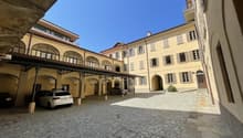 Cobblestone courtyard with cars parked under a covered area, yellow building with arched windows and doors, balconies, and a small plant in front of the entrance.