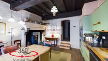 Rustic kitchen with exposed wooden beams, stone walls, and a fireplace. The kitchen features a wooden dining table, wicker chairs, and various kitchen appliances and utensils. There is also a 360 degree virtual tour sign visible.