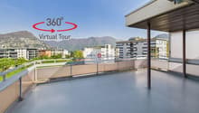 The image shows a 360-degree virtual tour of a balcony or terrace overlooking a city. The balcony has a concrete floor and a wooden roof structure. The view includes several multi-story buildings in the distance, with mountains visible in the background.