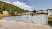 Garage building with multiple storage units, concrete floor, sign reading 'ABETE ROSSO Immobiliare', surrounded by greenery and a mountain in the background