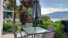 Outdoor patio with a glass-topped table and chairs, surrounded by lush tropical plants and flowers. There is a closed umbrella and a view of the mountains and lake in the background.